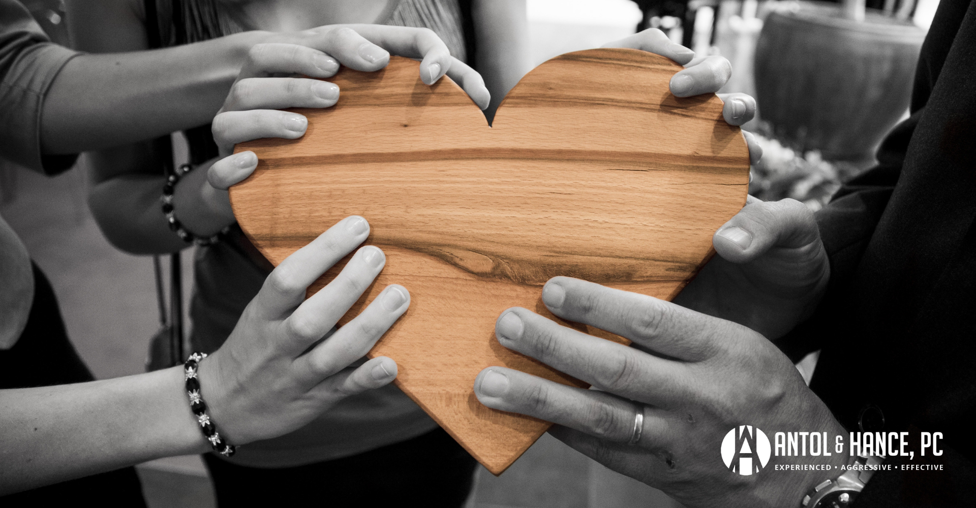 Image shows a group of people in shades of black and white placing their hands on a beige wooden heart.