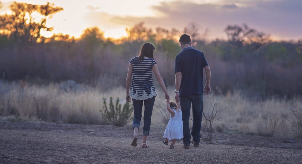 This image shows a family which includes a young mother, father, and toddler in a white dress, walking into the desert against a setting sun, facing the sunset. The mother, father, and child hold hands as a they walk away, with the child in between the two adults.