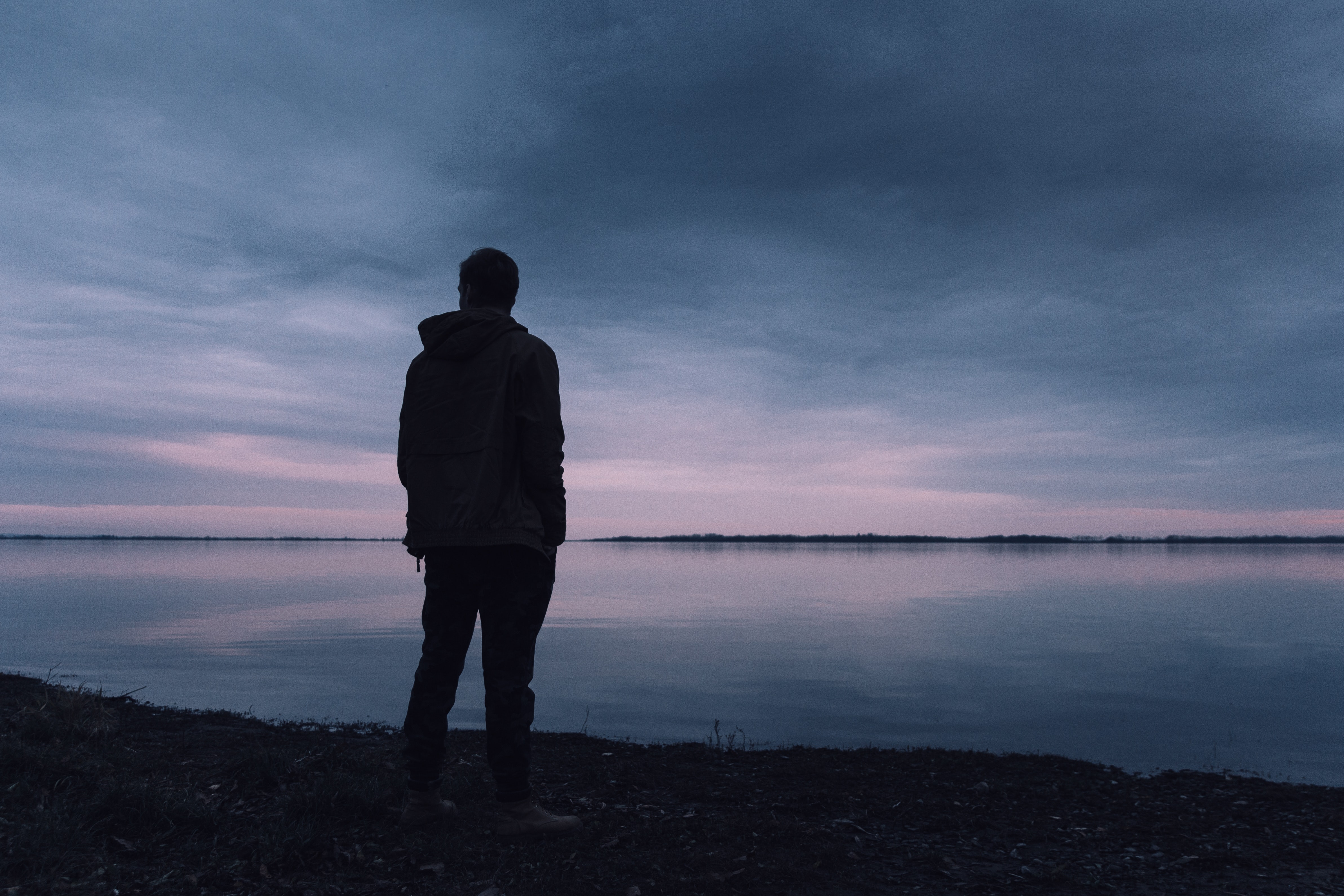 The dark silhouette of a man stands at the edge of a large glassy lake as the sun begins to rise in the distance and illuminate a cloudy sky.