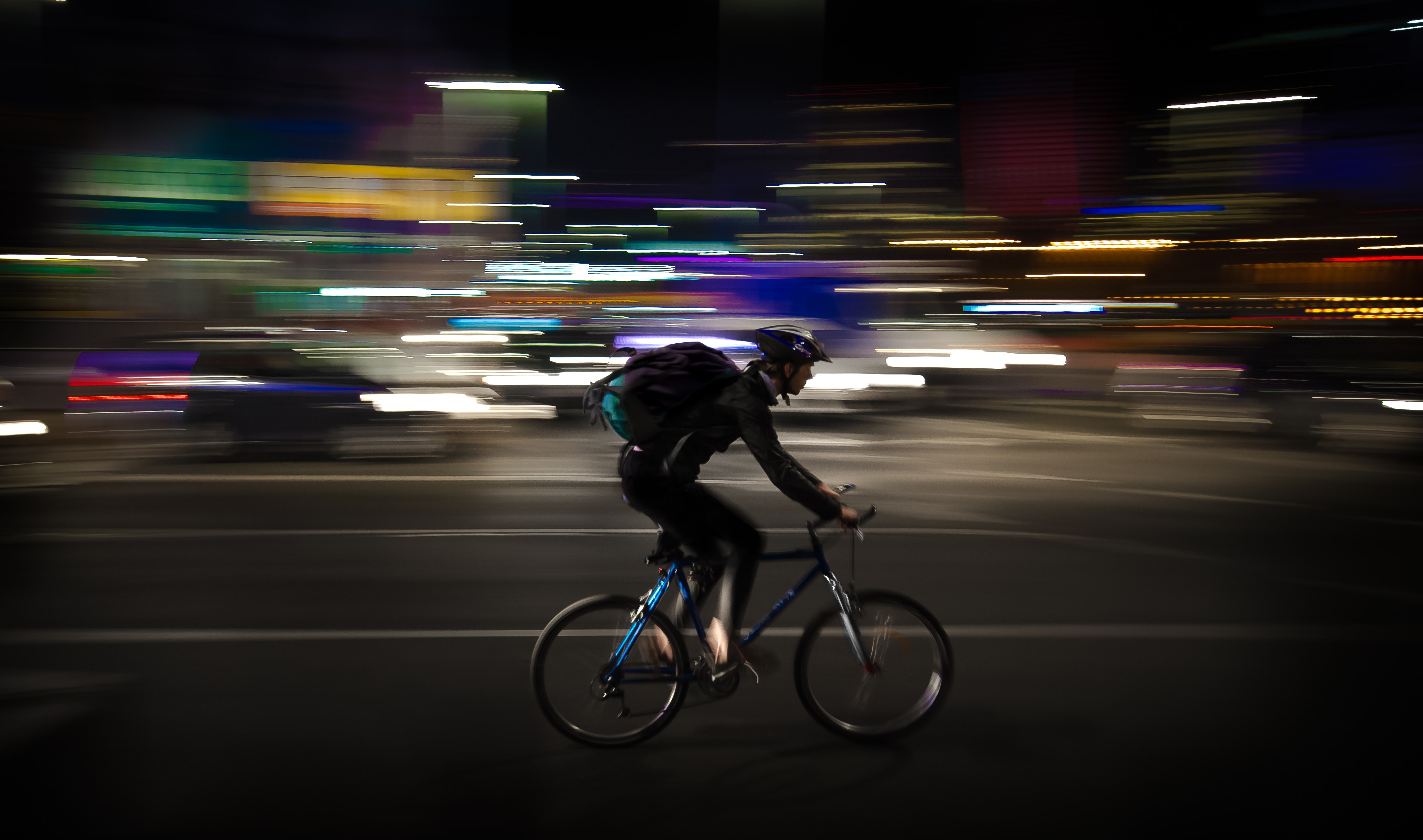 A man wearing a helmet and a green backpack rides his bike through a brightly lit city at night, the lights blurring and blending behind him as he rides along the street.
