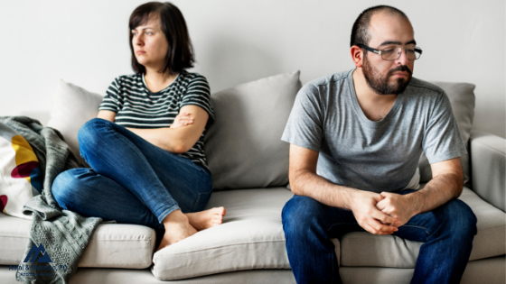 Man and woman sitting on a couch looking angry at each other. The woman is sitting on one end of the couch with her arms crossed and the man is sitting on the other end, slouched over. Both have disgruntled looks on their faces. 