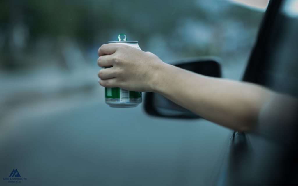 Someone behind the wheel in a vehicle, sticking their hand out of the window, firmly grasping a beer.