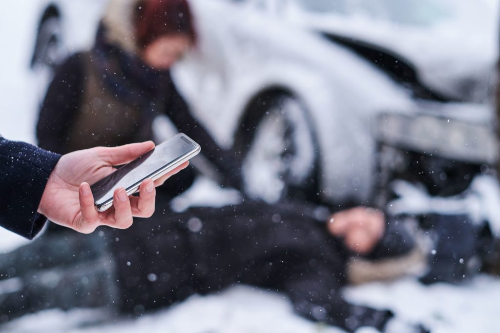 someone holding phone in front of the scene of a car accident with one person laying on the ground and another tending to them