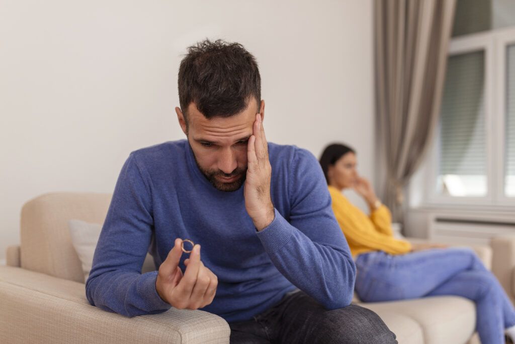 stressed man touching his face and looking at his wedding ring with a spouse on the other side of the couch