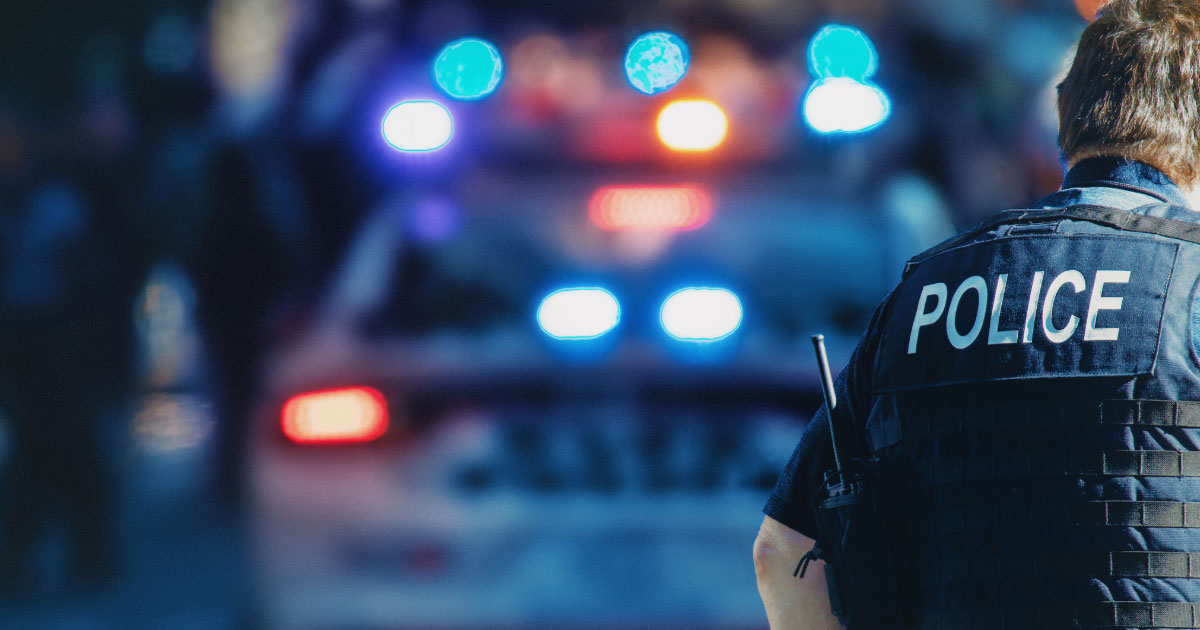 Police officer wearing vest standing in front of cop car with lights on